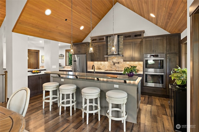 kitchen featuring hanging light fixtures, a kitchen island with sink, stainless steel appliances, wall chimney range hood, and dark brown cabinetry