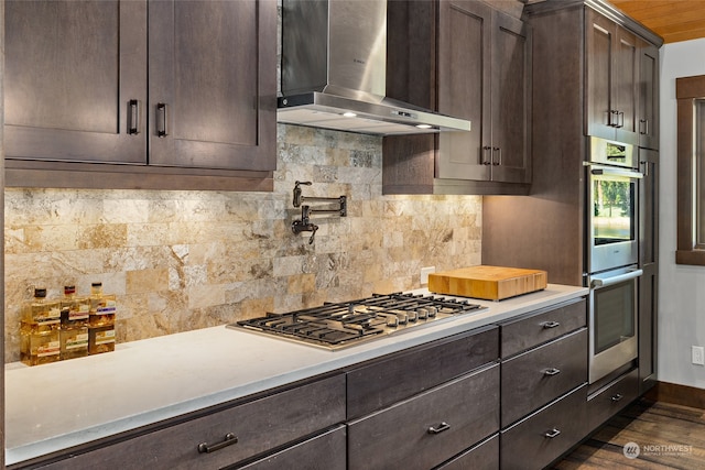 kitchen featuring dark hardwood / wood-style floors, stainless steel appliances, wall chimney range hood, dark brown cabinets, and tasteful backsplash