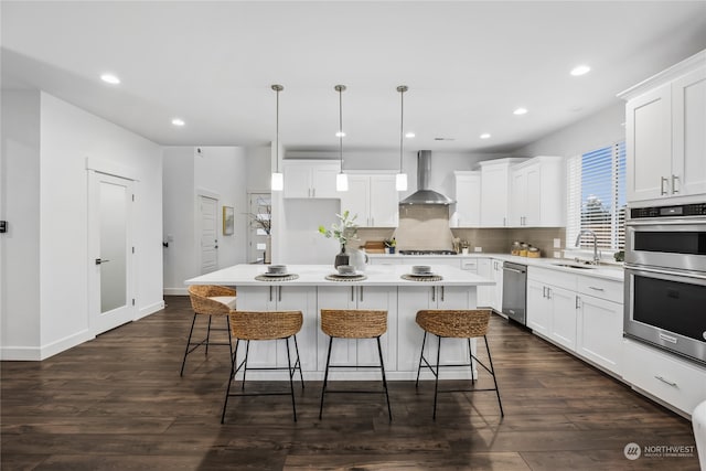 kitchen with white cabinetry, hanging light fixtures, a kitchen island, stainless steel appliances, and wall chimney range hood