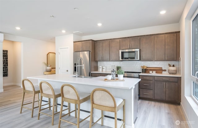 kitchen with a kitchen island with sink, backsplash, light wood-type flooring, stainless steel appliances, and a breakfast bar area