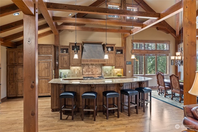 kitchen with a large island with sink, hanging light fixtures, light stone countertops, light wood-type flooring, and custom range hood