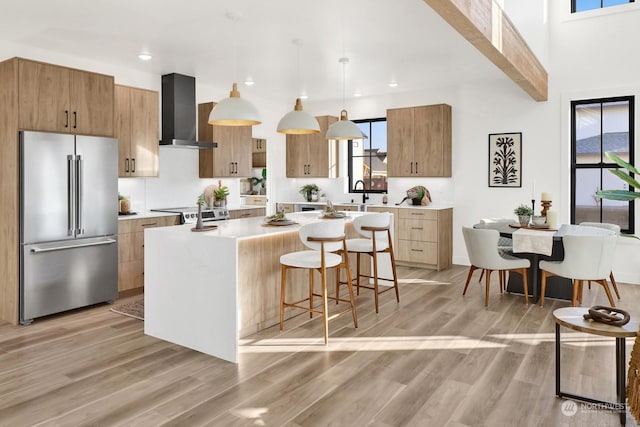 kitchen with an island with sink, high end refrigerator, hanging light fixtures, wall chimney range hood, and light hardwood / wood-style flooring