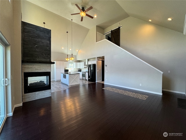 unfurnished living room featuring high vaulted ceiling, a barn door, ceiling fan, a multi sided fireplace, and hardwood / wood-style flooring