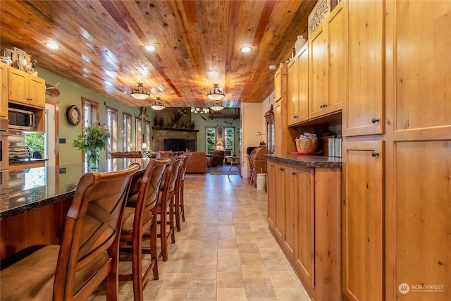 kitchen featuring a large fireplace, wooden ceiling, stainless steel microwave, light tile floors, and dark stone countertops