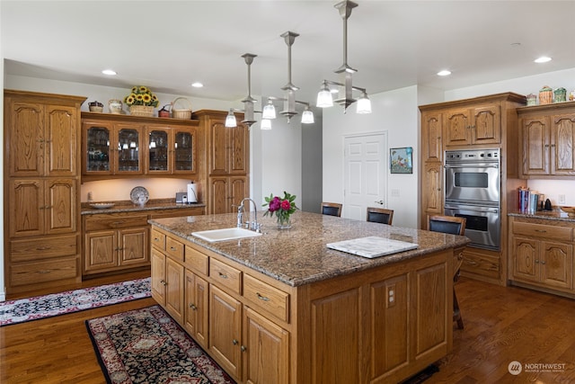 kitchen featuring double oven, dark hardwood / wood-style flooring, a kitchen island with sink, sink, and pendant lighting