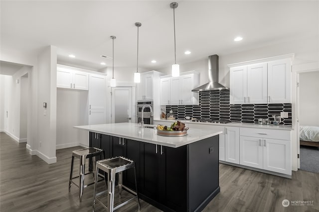 kitchen with white cabinets, pendant lighting, wall chimney exhaust hood, and an island with sink