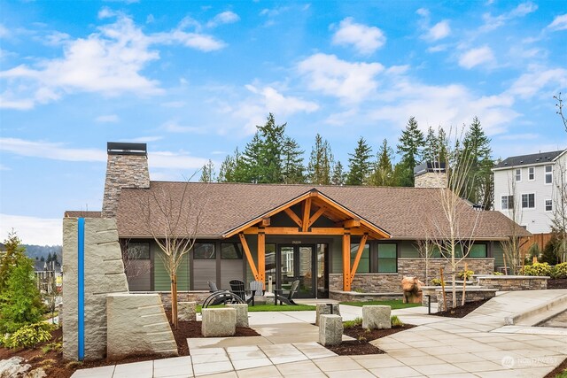 exterior space with a patio, stone siding, concrete driveway, roof with shingles, and a chimney