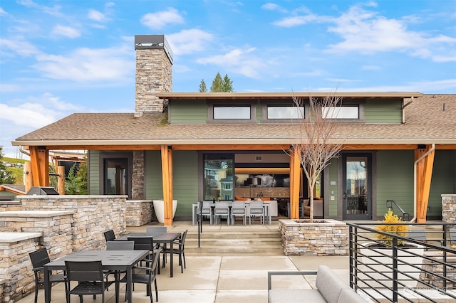 rear view of property featuring outdoor dining area, a shingled roof, exterior kitchen, a chimney, and a patio area