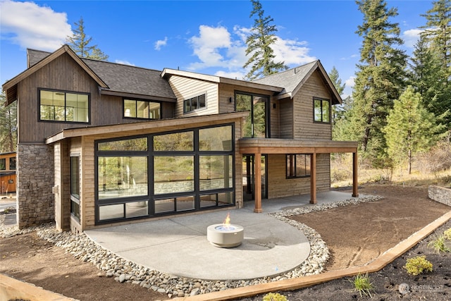 rear view of house featuring an outdoor fire pit, stone siding, roof with shingles, and a patio