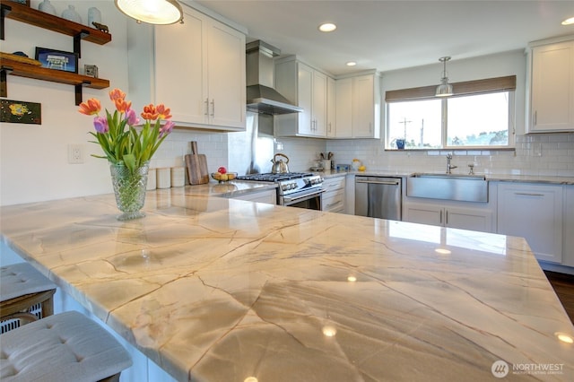 kitchen featuring a sink, appliances with stainless steel finishes, wall chimney range hood, and light stone counters