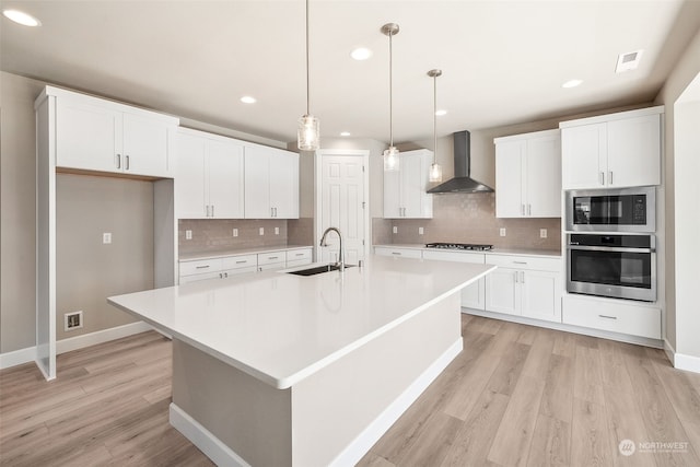 kitchen featuring light wood-style flooring, appliances with stainless steel finishes, wall chimney range hood, white cabinetry, and a sink