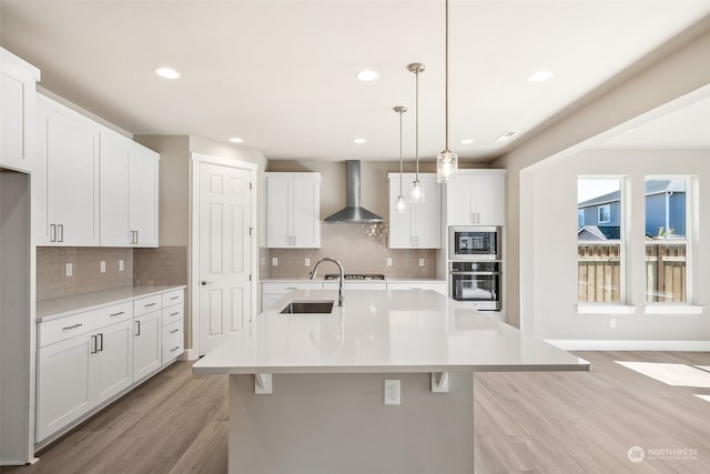 kitchen featuring a center island with sink, appliances with stainless steel finishes, light countertops, wall chimney range hood, and a sink