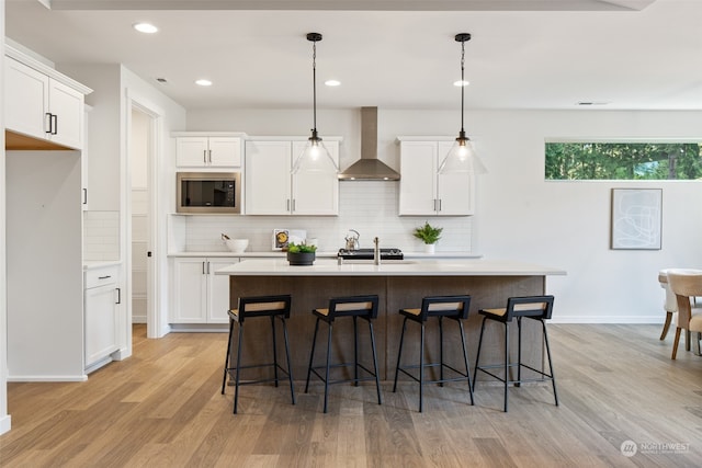 kitchen with built in microwave, wall chimney exhaust hood, an island with sink, pendant lighting, and light wood-type flooring