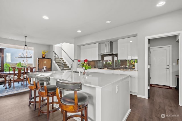 kitchen with dark hardwood / wood-style flooring, wall chimney range hood, decorative light fixtures, a center island with sink, and white cabinetry