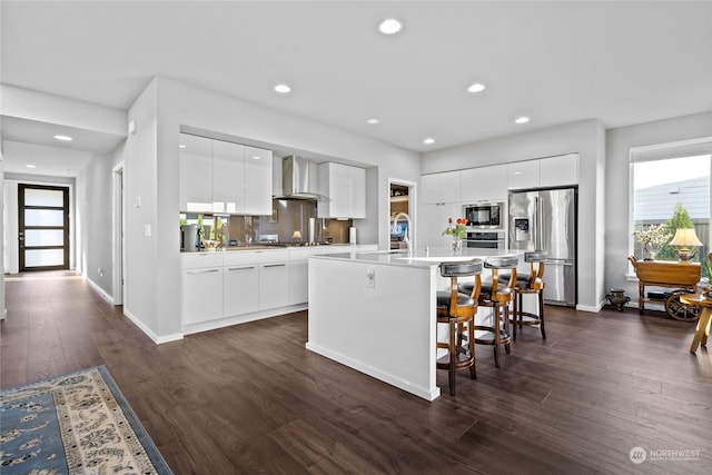 kitchen featuring appliances with stainless steel finishes, dark hardwood / wood-style flooring, a kitchen island with sink, wall chimney range hood, and white cabinets