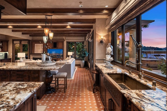 kitchen with light stone countertops, decorative light fixtures, beam ceiling, a breakfast bar area, and dark brown cabinets