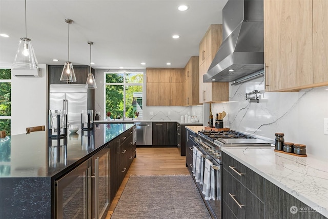 kitchen with hanging light fixtures, wall chimney exhaust hood, dark stone countertops, appliances with stainless steel finishes, and light hardwood / wood-style floors