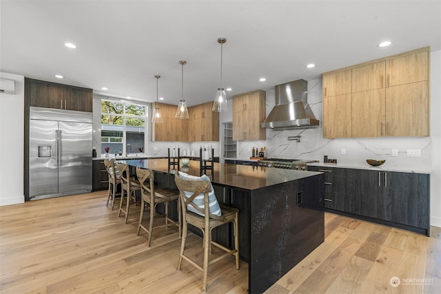 kitchen featuring light hardwood / wood-style floors, stainless steel built in refrigerator, wall chimney range hood, and a kitchen island with sink