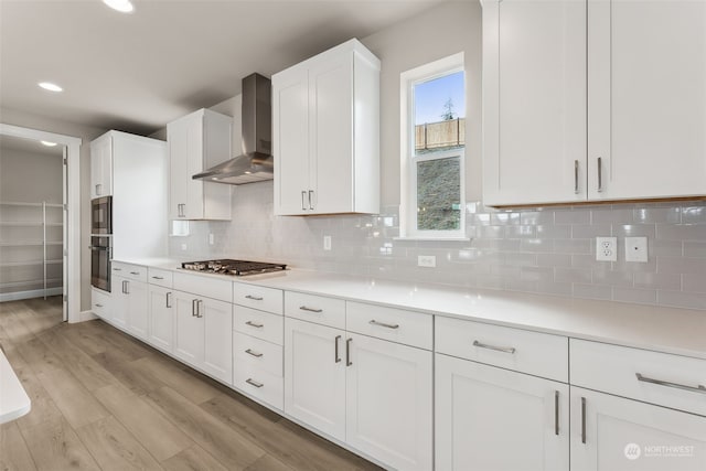 kitchen featuring light wood-type flooring, tasteful backsplash, wall chimney exhaust hood, white cabinetry, and appliances with stainless steel finishes