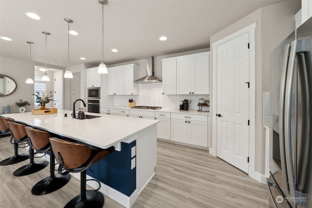 kitchen featuring appliances with stainless steel finishes, hanging light fixtures, an island with sink, white cabinets, and wall chimney range hood