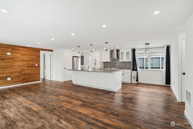 kitchen featuring pendant lighting, a kitchen island with sink, wall chimney range hood, stainless steel fridge, and white cabinetry