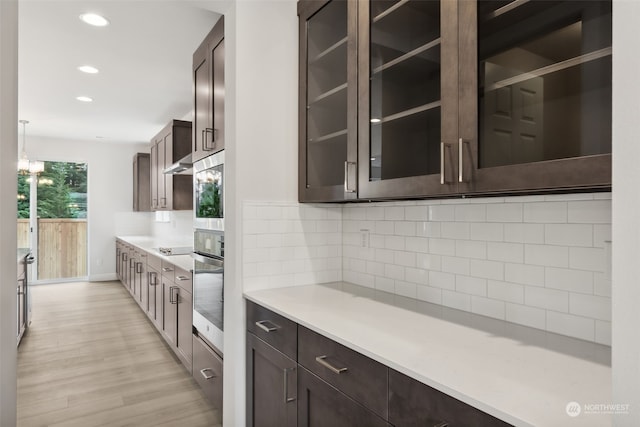 kitchen featuring stainless steel oven, hanging light fixtures, dark brown cabinetry, light wood-type flooring, and tasteful backsplash