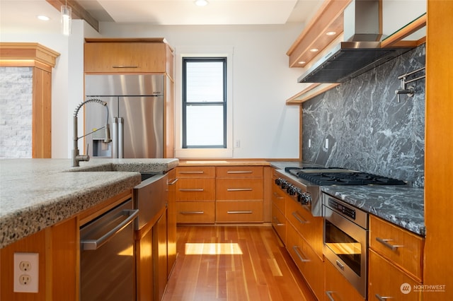 kitchen with light stone countertops, appliances with stainless steel finishes, wall chimney exhaust hood, hardwood / wood-style floors, and tasteful backsplash