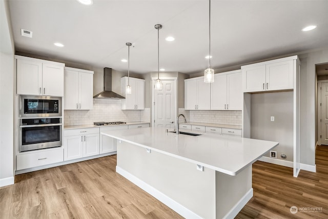 kitchen with wall chimney range hood, sink, white cabinetry, stainless steel appliances, and a center island with sink