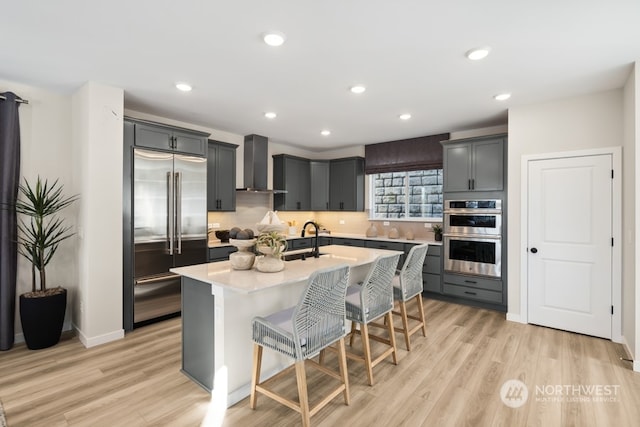 kitchen featuring wall chimney range hood, appliances with stainless steel finishes, light hardwood / wood-style floors, an island with sink, and a kitchen bar