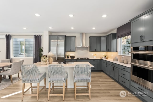 kitchen featuring a breakfast bar, gray cabinetry, stainless steel appliances, wall chimney exhaust hood, and light wood-type flooring