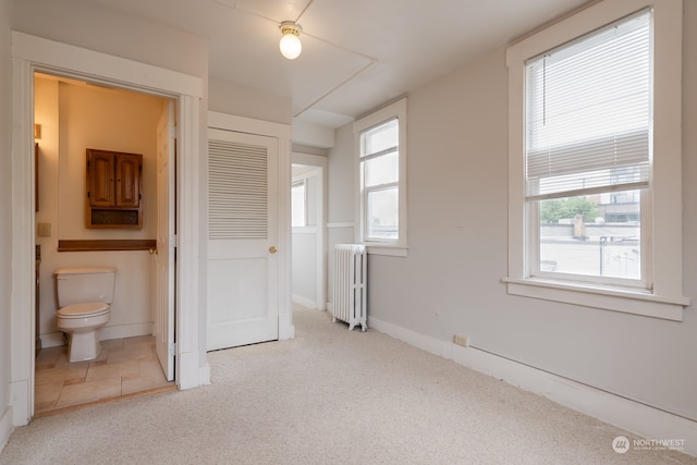 unfurnished bedroom with a closet, ensuite bathroom, ceiling fan, radiator, and light colored carpet