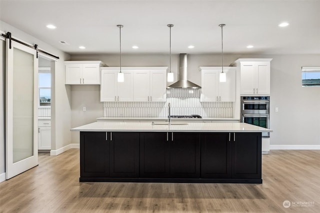 kitchen with a barn door, white cabinets, wall chimney exhaust hood, and an island with sink