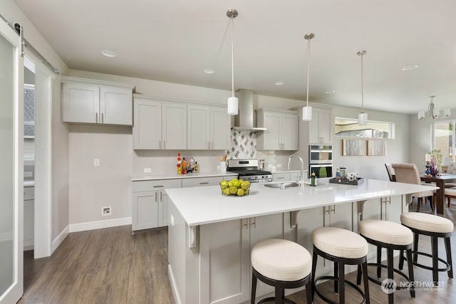 kitchen with pendant lighting, a center island with sink, wall chimney range hood, a barn door, and stainless steel appliances