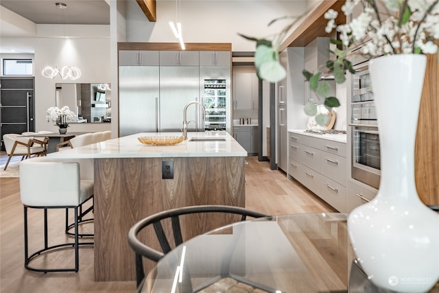 kitchen with stainless steel oven, decorative light fixtures, an island with sink, sink, and light hardwood / wood-style flooring