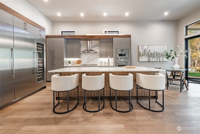 kitchen with light wood-type flooring, wall chimney exhaust hood, a center island with sink, appliances with stainless steel finishes, and gray cabinetry