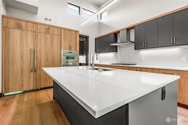 kitchen with a sink, wall chimney exhaust hood, appliances with stainless steel finishes, visible vents, and a towering ceiling