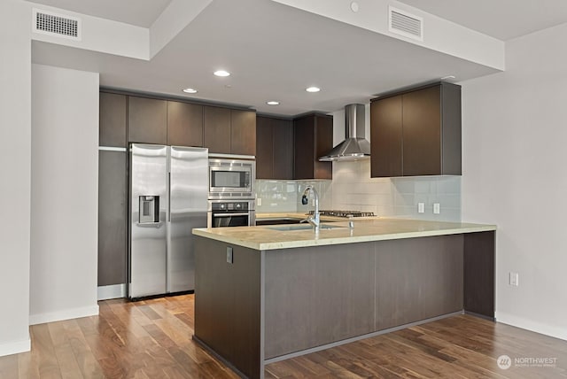 kitchen featuring wall chimney exhaust hood, dark brown cabinetry, sink, appliances with stainless steel finishes, and kitchen peninsula