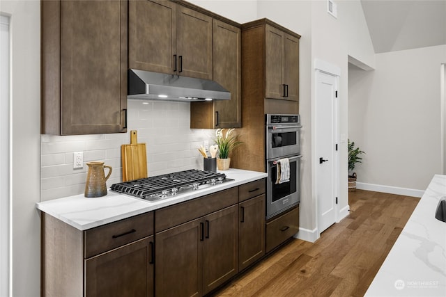 kitchen with appliances with stainless steel finishes, under cabinet range hood, light stone countertops, wood finished floors, and backsplash