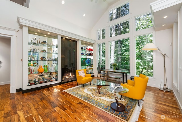 living area with high vaulted ceiling, a glass covered fireplace, wood-type flooring, and baseboards