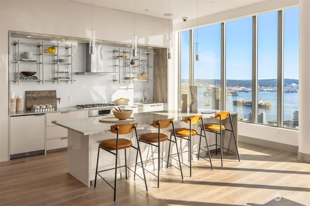 bar featuring wall chimney range hood, white cabinetry, a water view, tasteful backsplash, and decorative light fixtures