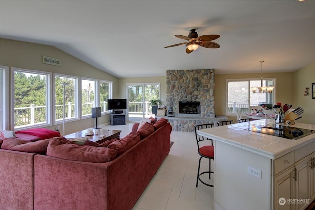 living room with ceiling fan with notable chandelier, a wealth of natural light, lofted ceiling, and a stone fireplace