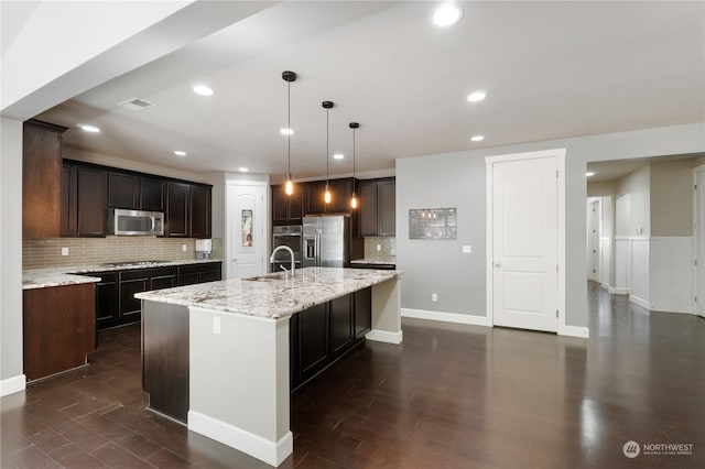 kitchen with tasteful backsplash, a kitchen island with sink, light stone countertops, appliances with stainless steel finishes, and dark wood-type flooring