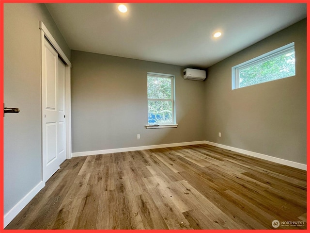 unfurnished bedroom with multiple windows, a wall mounted air conditioner, baseboards, and wood finished floors