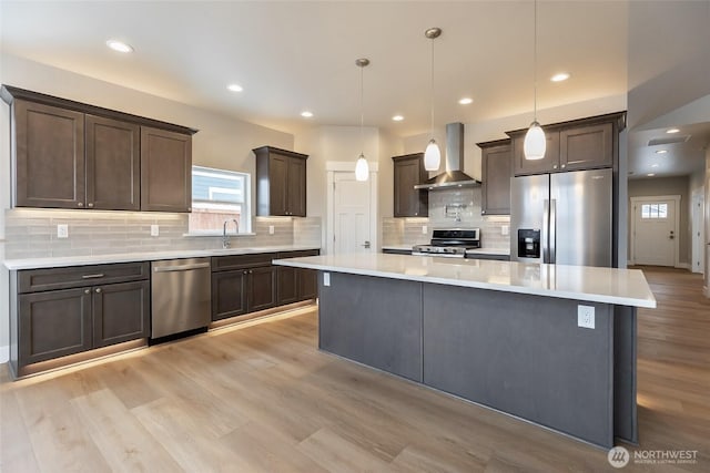 kitchen with a kitchen island, light countertops, appliances with stainless steel finishes, hanging light fixtures, and wall chimney range hood