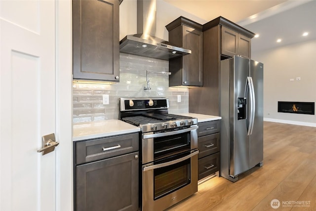 kitchen featuring light wood-style flooring, stainless steel appliances, dark brown cabinets, light countertops, and wall chimney exhaust hood