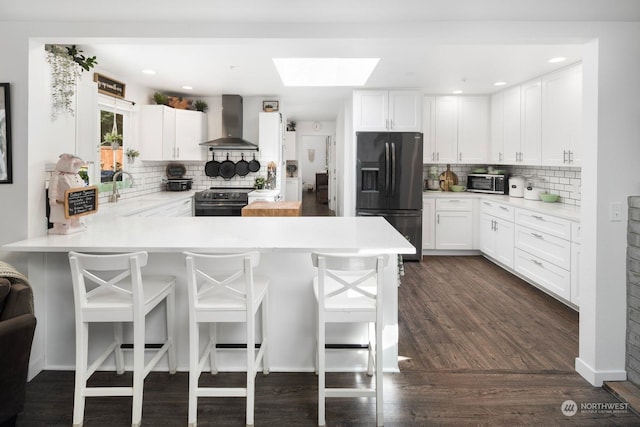 kitchen featuring kitchen peninsula, black fridge with ice dispenser, wall chimney range hood, white cabinetry, and a breakfast bar area