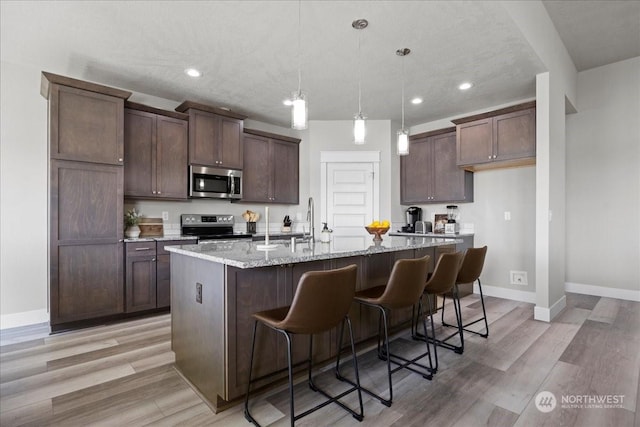 kitchen with baseboards, an island with sink, light wood-style flooring, stainless steel appliances, and a sink