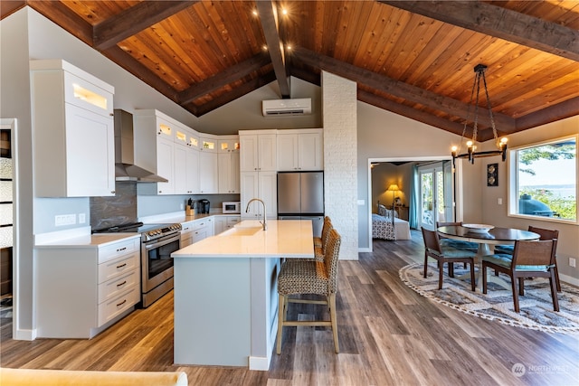 kitchen with a wall mounted AC, a center island with sink, wall chimney range hood, stainless steel appliances, and white cabinets