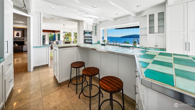 kitchen featuring oven, tile countertops, beam ceiling, white cabinetry, and stainless steel microwave