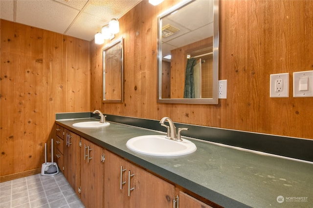 bathroom featuring tile patterned floors, a drop ceiling, wooden walls, and dual bowl vanity